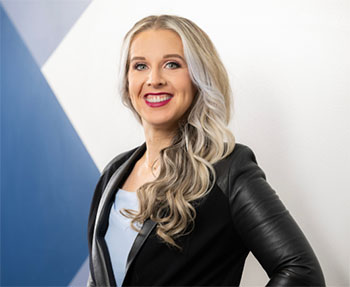 Professional headshot of Sarah Arten with long blonde hair, wearing a black blazer and light top against a modern geometric background