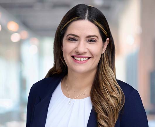 Professional headshot of Teresa Martinez with long dark hair, wearing a navy blazer and white blouse in a softly blurred office hallway setting