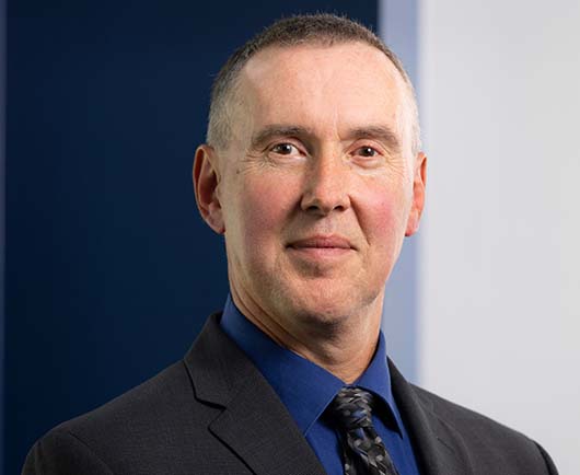 Professional headshot of Terry Thiem with short hair, wearing a dark suit, blue shirt, and patterned tie against a modern blue and white background