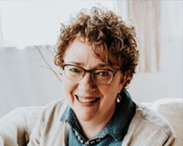Professional headshot of Tami Thompson with short curly hair and glasses, wearing a casual layered outfit in a softly lit home or office setting