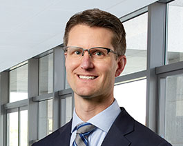Professional headshot of Tom Mueller wearing glasses, a suit, and striped tie, standing in a modern office with large windows