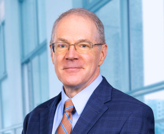 Professional headshot of William Quigley with glasses, wearing a blue suit and orange striped tie, standing in front of large office windows