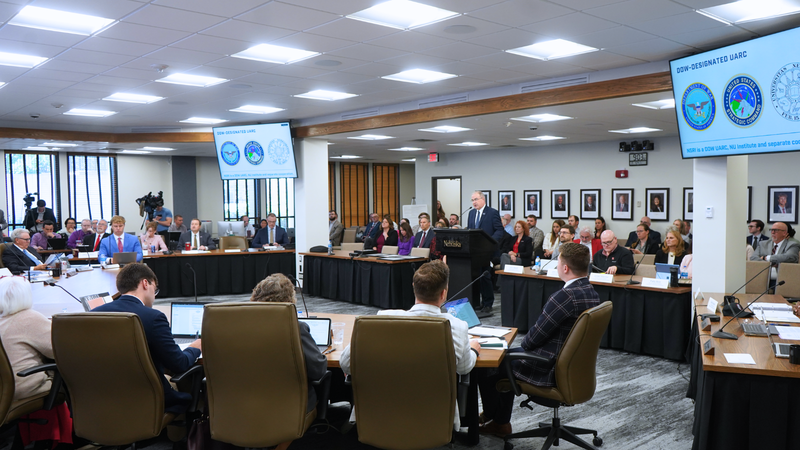 Presenter addressing Board of Regents meeting in large conference room with attendees seated at U-shaped table, organizational logos displayed on wall-mounted screens