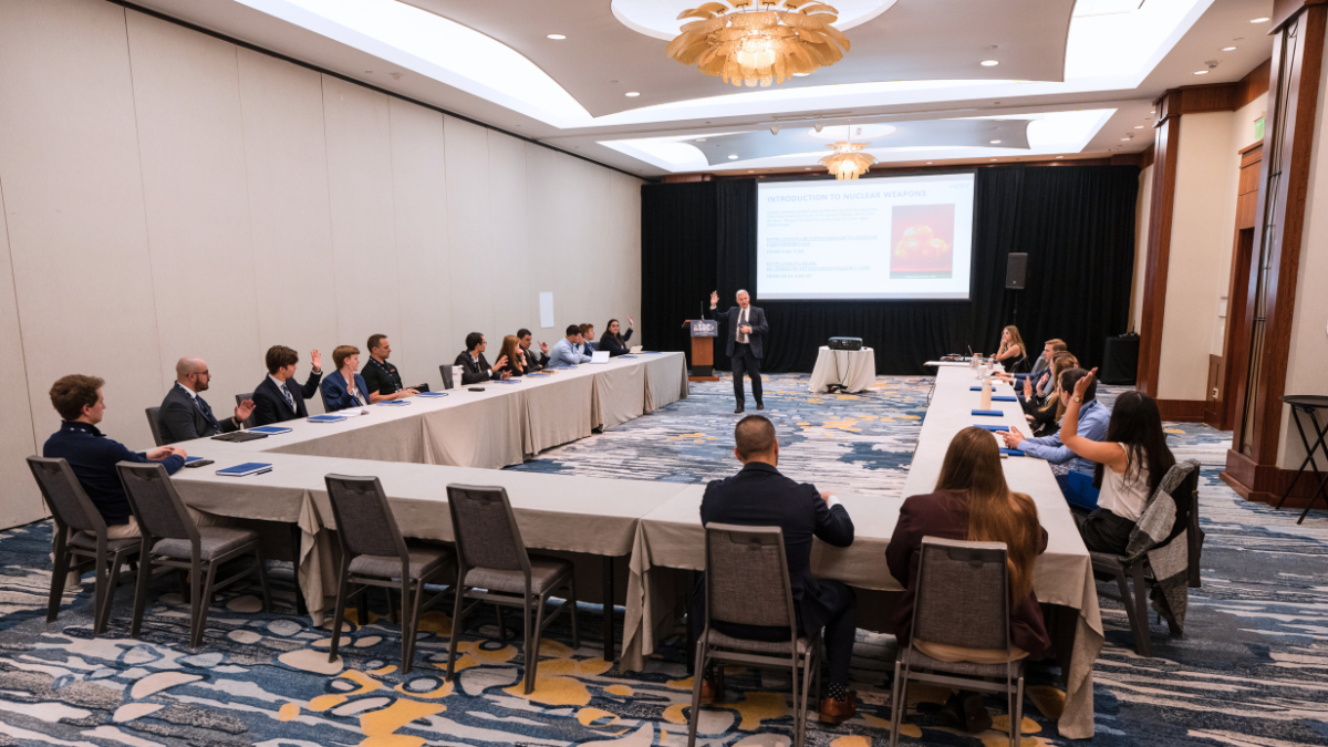 Pat Rhoads presents to a group of young professionals seated at a U-shaped table arrangement in a hotel conference room. Several attendees raise their hands in response to a question. A projected slide titled