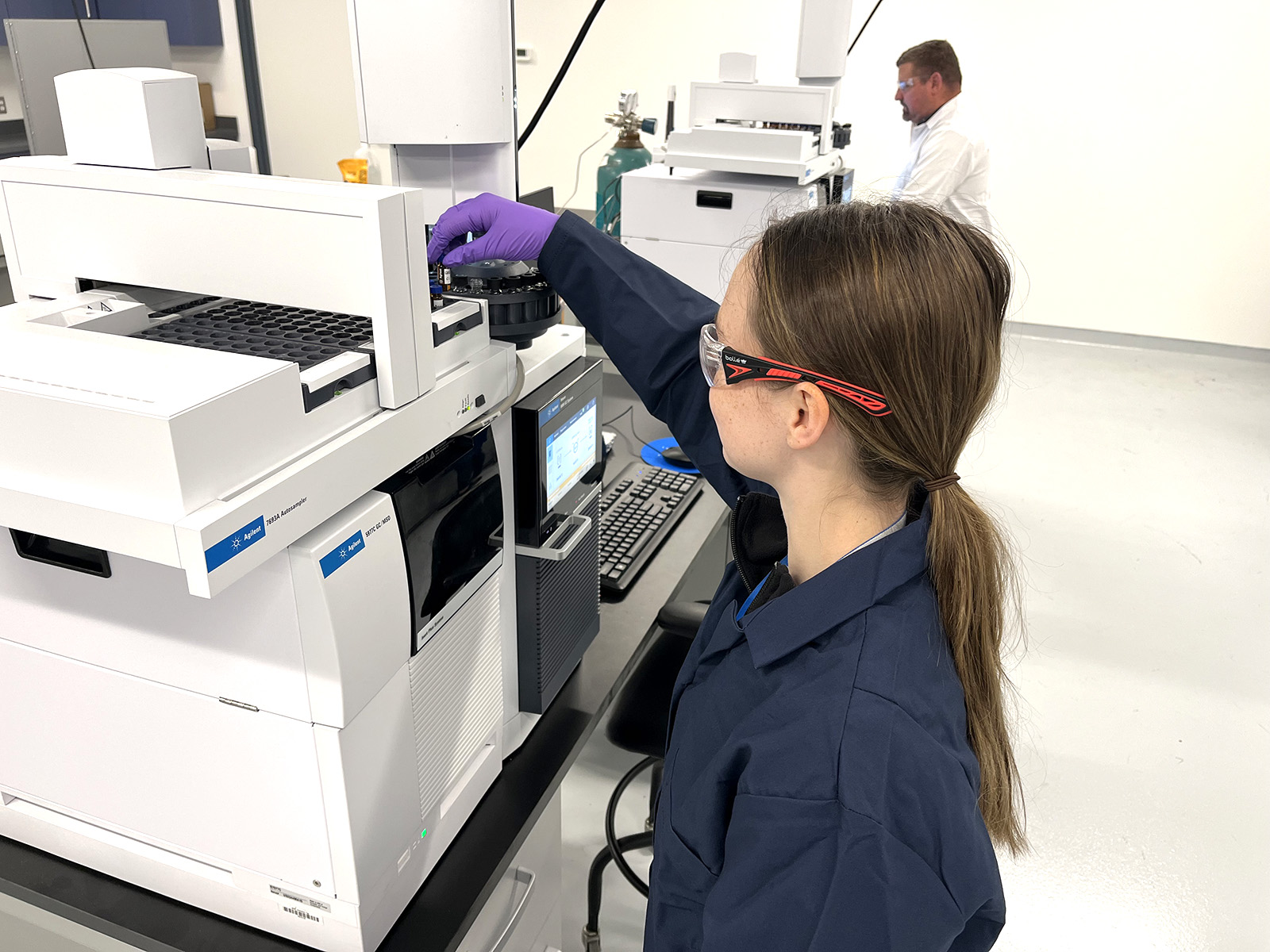 Two individuals working in a laboratory with advanced analytical equipment. One individual is operating a large Agilent instrument, wearing a lab coat, safety glasses, and gloves, while inserting a sample into the machine. The lab features clean white surfaces, computer monitors, gas cylinders, and additional scientific instruments in the background.