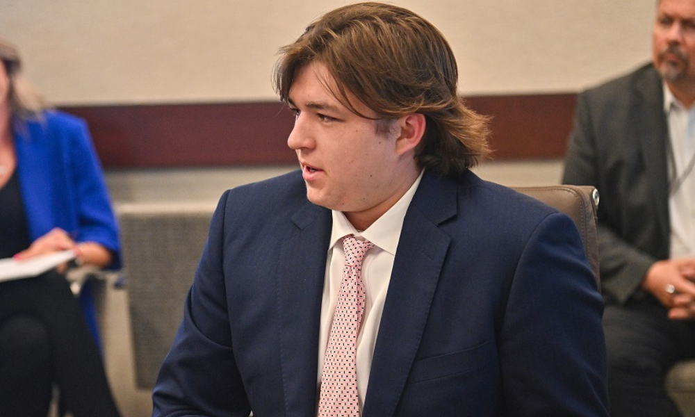 Young male student in suit sitting at conference table talking to someone off camera