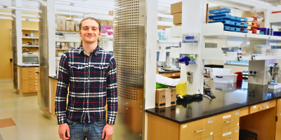 Two individuals sit at a table in a lab, examining scientific equipment. The table displays various lab items, including a pipette and test tubes. In the background, there is a large poster and lab equipment.
